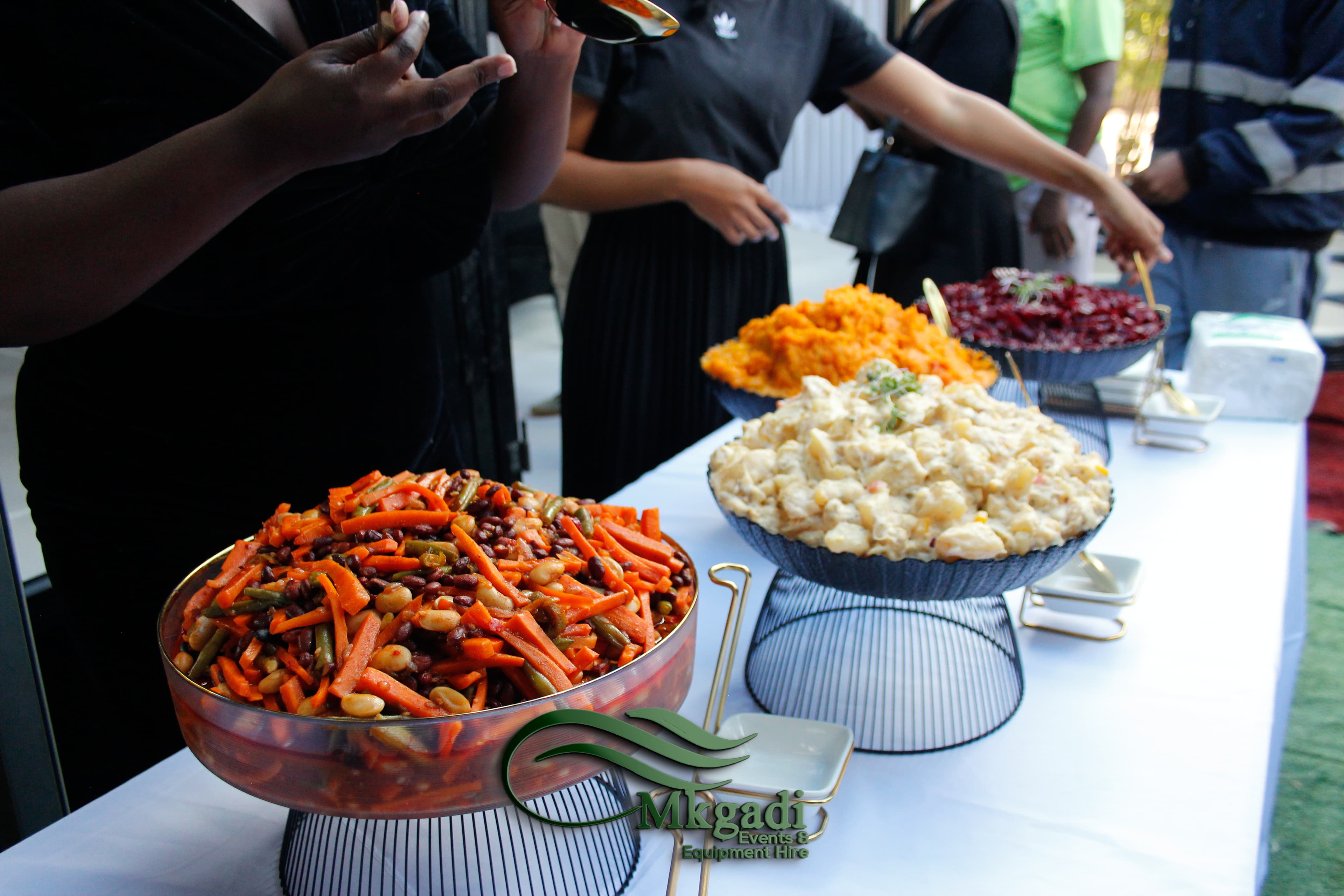 Outdoor funeral catering buffet with colourful salad bowls on a white linen table, guests in black attire serving themselves